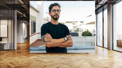 Confident stylish guy with tattoos posing on apartment balcony or terrace. Young man in glasses standing outside with arms crossed and looking at camera. Male portrait concept Wall mural