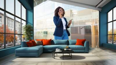 Content businesswoman walking and talking by smartphone. Cheerful young African American businesswoman talking by cell phone and looking away. Communication concept Wall mural