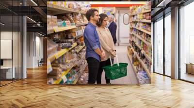 Couple looking at shelves in grocery store. Focused young man and woman holding basket and choosing products in supermarket. Shopping concept Wall mural