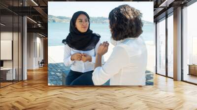 Diverse female coworkers relaxing and chatting outside. Young Muslim businesswoman in black hijab sitting on promenade near river and talking to female colleague. Office friends concept Wall mural