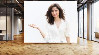 Doubtful woman pointing hand at empty copy space and shrugging. Wavy haired young woman in casual shirt standing isolated over white background. Advertising concept Wall mural