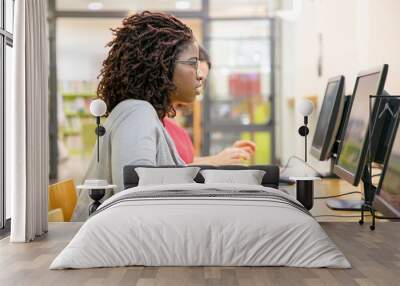 Focused female student excited with online test in computer class. Black woman in casual sitting at table, using desktop, typing, looking at monitor with open mouth. Excites student concept Wall mural