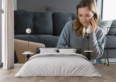 Focused freelancer writing down customer instructions. Young woman in casual sitting at laptop in her living room, talking on phone and writing notes. Multitasking concept Wall mural