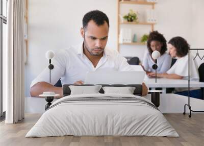 Focused male employee using laptop in office. His multiethnic colleagues working together and talking in background. Sharing office concept Wall mural