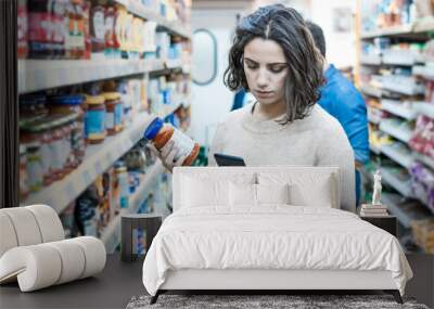 Focused woman using smartphone in grocery store. Young woman reading checklist via smartphone and choosing products in grocery store. Supermarket concept Wall mural