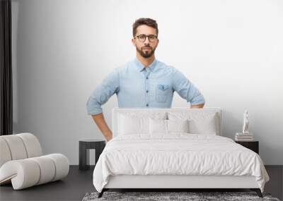 Front of serious determined guy keeping hands on hip. Handsome young man in casual shirt and glasses standing isolated over white background. Male portrait concept Wall mural