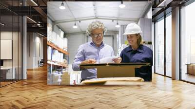 Grey haired mature professional and female worker in hardhat talking and reviewing documents in warehouse. Front view. Labor or safety inspection concept Wall mural