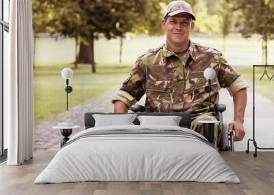 Happy disabled military man in wheelchair wearing camouflage uniform, moving on footpath in city park. Front view. Veteran of war or disability concept Wall mural