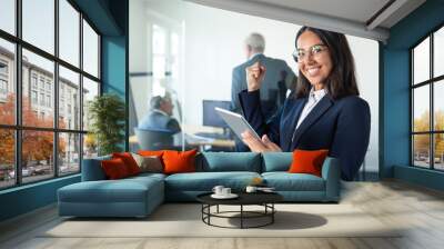 Happy female professional in glasses and suit holding tablet and making winner gesture while two businessmen working behind glass wall. Copy space. Communication concept Wall mural