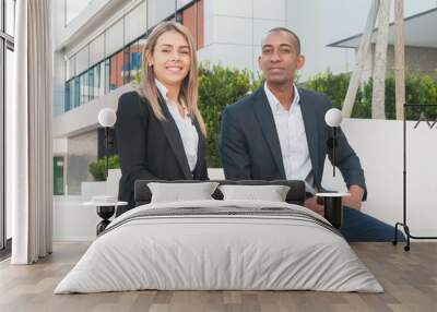 Happy successful business people posing outdoors. Black man and Caucasian woman in office suits sitting outside, looking at camera and smiling. Business team concept Wall mural