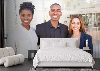 Happy united multiethnic team posing in office hall. Three professionals in formal suits standing for camera indoors and smiling. Corporate friendship concept Wall mural