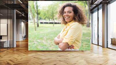 Happy woman keeping arms crossed and posing at camera in park. Young woman looking at camera with blurred green trees and lawn in background. Nature and leisure concept. Side view. Wall mural