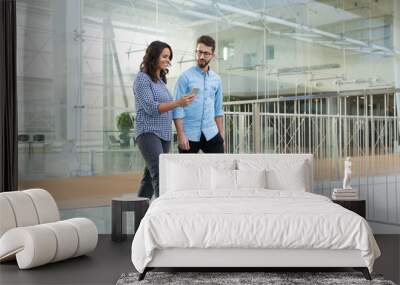 Joyful colleagues in casual looking at cellphone screen together while going through office hallway. Young man and woman walking indoors with glass wall in background. Communication concept Wall mural