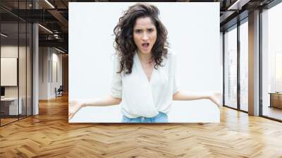 Mad exaggerated woman gasping, spreading hands and asking question. Wavy haired young woman in casual shirt standing isolated over white background. Conflict concept Wall mural