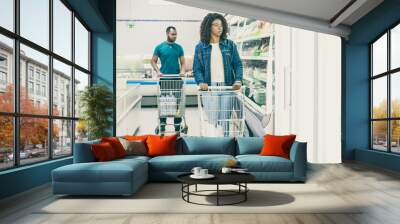 Multiethnic customers walking along isles in grocery store, wheeling carts, choosing product. Buyers shopping in supermarket. Buying fresh food concept Wall mural