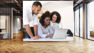 Multiethnic team collaborating on project at one workplace. Business man and women in casual sitting and standing at desk, using laptop, looking at screen and talking. Collaboration concept Wall mural