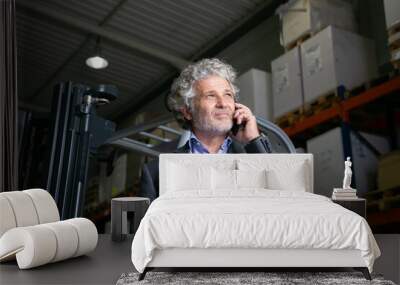 Pensive mature businessman standing near forklift in warehouse and speaking on cell phone. Shelves with goods in background. Business or logistics concept Wall mural