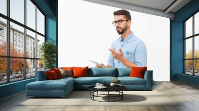 Pensive tablet user looking at camera and pointing finger away. Handsome young man in casual shirt and glasses standing isolated over white background. Advertising concept Wall mural