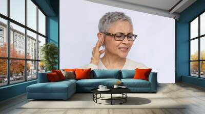 Portrait of irritated senior woman suffering from eardrum. Mature Caucasian woman wearing eyeglasses and white jumper touching ear, annoyed with noise. Earache concept Wall mural