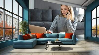 Positive successful businesswoman enjoying work on weekend. Young woman in casual using laptop and talking on phone in her living room. Business at home concept Wall mural