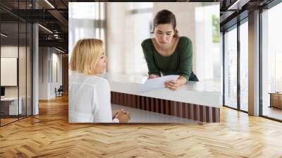 Puzzled female guest having trouble with filling out hotel documents and consulting receptionist. Young woman leaning on reception desk and pointing pen at paper. Business hotel concept Wall mural