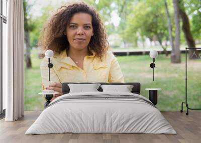 Relaxed woman keeping arms crossed and posing at camera in park. Young woman looking at camera with blurred green trees and lawn in background. Nature and leisure concept. Side view. Wall mural