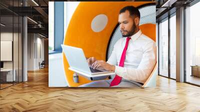 Serious businessman using laptop computer. Focused young African American businessman working with laptop in modern office. Business and technology concept Wall mural