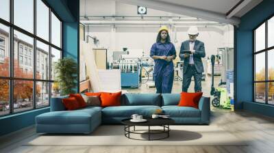 Serious male inspector and black female factory employee in hardhats walking on plant floor and talking, man using tablet. Front view, full length. Labor and production concept Wall mural