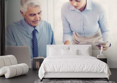 Serious senior business man and female secretary are discussing document. Woman is pointing to it and bending over desk. Man is sitting at desk near her in office with blurred window in background. Wall mural