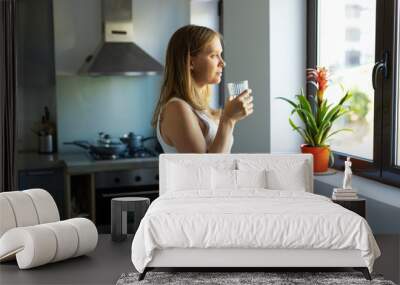 Side of pensive expectant mother standing in kitchen with glass of water, looking through window. Pregnant young woman spending leisure time at home. Hydration and prenatal care concept Wall mural