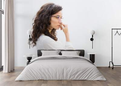 Side of pensive office assistant in glasses leaning face on hand and looking away at copy space. Wavy haired young woman in casual shirt standing isolated over white background. Advertising concept Wall mural