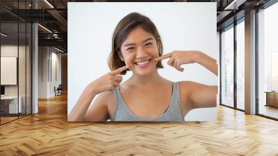 Smiling pretty Asian woman pointing at her teeth. Young woman showing teeth and looking at camera. Teeth hygiene and whitening concept. Isolated front view on white background. Wall mural