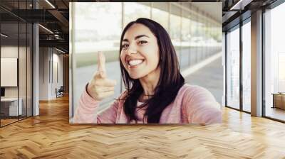 Smiling woman taking selfie photo and pointing at you outdoors. Pretty young lady looking at camera with building wall and walkway in background. Selfie and tourism concept. Front view. Wall mural