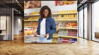 Smiling young customer choosing goods. Cheerful African American woman walking with shopping cart in supermarket. Shopping concept Wall mural