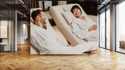 Smiling young woman and man wearing bathrobes, chatting and lying on deckchairs in spa salon Wall mural