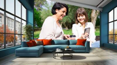 Smiling young woman showing smartphone screen to mother in park. Middle-aged lady and her daughter sitting on bench and relaxing with trees in background. Family and communication concept. Front view. Wall mural