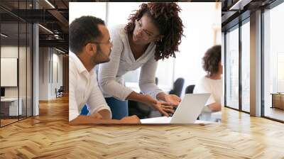 Trainer explaining software specifics to new employee. Man and woman in casual sitting and standing at desk, using laptop, pointing at screen and talking. Corporate education concept Wall mural