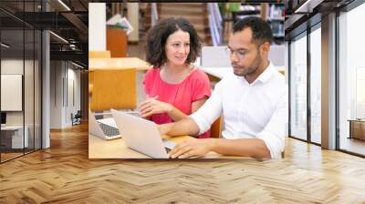 Two adult college students chatting while working in library computer class. Man and woman in casual sitting at desk, using laptops and talking. Communication concept Wall mural