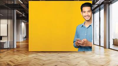 Asian man holding mobile phone smiling while standing isolated on yellow background with copy space for text, People on smart phone, technology lifestyle Wall mural