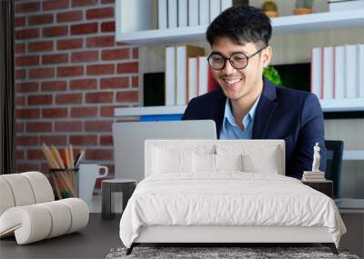 Young asian businessman smiling while working with laptop computer at office, business office lifestyle concept Wall mural