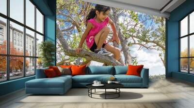 A young schoolgirl enjoys being high up in the trees. On a beautiful sunny day at the park filled with small trees, an adventurous little girl makes her way up the tree. Wall mural