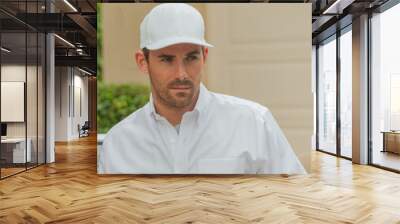 Closeup photo of a handsome entrepreneur wearing a white baseball cap and a white button-down shirt. The man looks away to his left focused on something in the distance. Wall mural