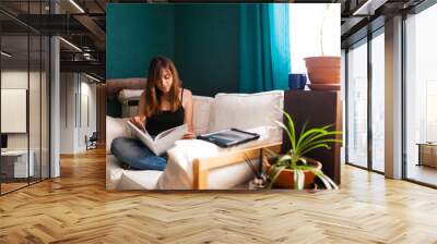 Young red-haired woman, reading a book in a couch surrounded by plants. Leisure indoors entertainment. Wall mural