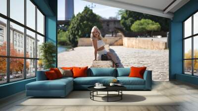 Beautiful young woman, blonde with blue eyes and wearing a white sequined dress, sitting on a stone wall, posing while on vacation in the city, with the Triana bridge in the background. Wall mural