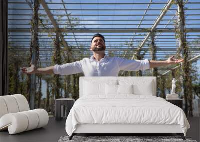 Handsome young man with beard, wearing white shirt and jeans, with open arms, very happy and grateful, looking at the sky. Concept happiness, success, gratitude, thanks to god. Wall mural