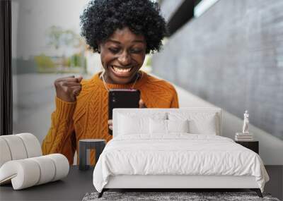 Image of a happy and emotional African American woman posing isolated on out of focus background using cell phone making a winning gesture. Wall mural