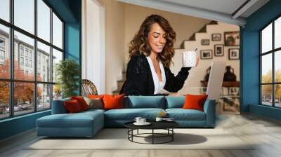 Portrait of a cheerful woman at home with a laptop and a cup of coffee on the table. 30 year old girl connected online to a meeting with friends and family. Telework concept Wall mural