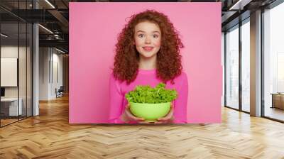 Smiling woman with curly hair holds a bowl of fresh green salad against a vibrant pink background, promoting healthy eating. Wall mural