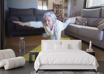 Older caucasian woman in sportswear doing gymnastic exercises on the living room floor with a mat Wall mural