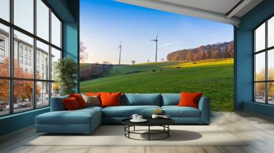 landscape cows grazing on grass field hill edge of forest and two tall wind turbines Wall mural